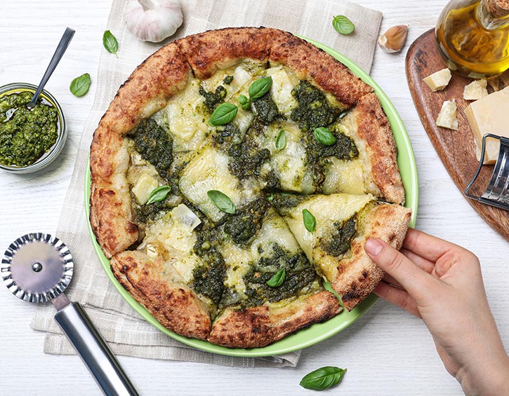 Hand lifting a slice of pesto pizza with cheese and basil from a green plate, surrounded by fresh ingredients and kitchen tools.