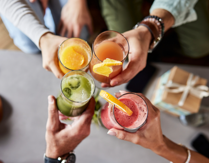 Group of people toasting with colorful drinks garnished with orange slices and mint, around a casual table with a wrapped gift.
